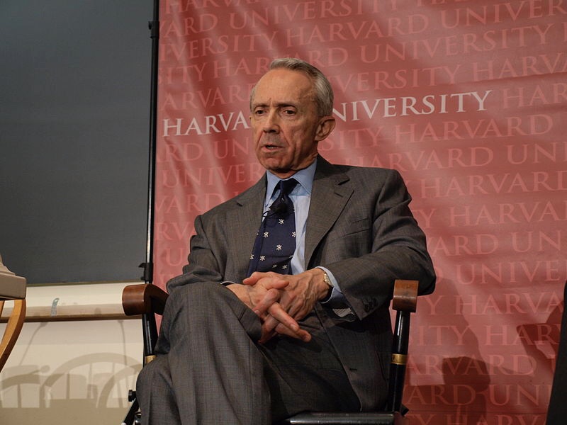 man in suit sitting in front of Harvard University banner
