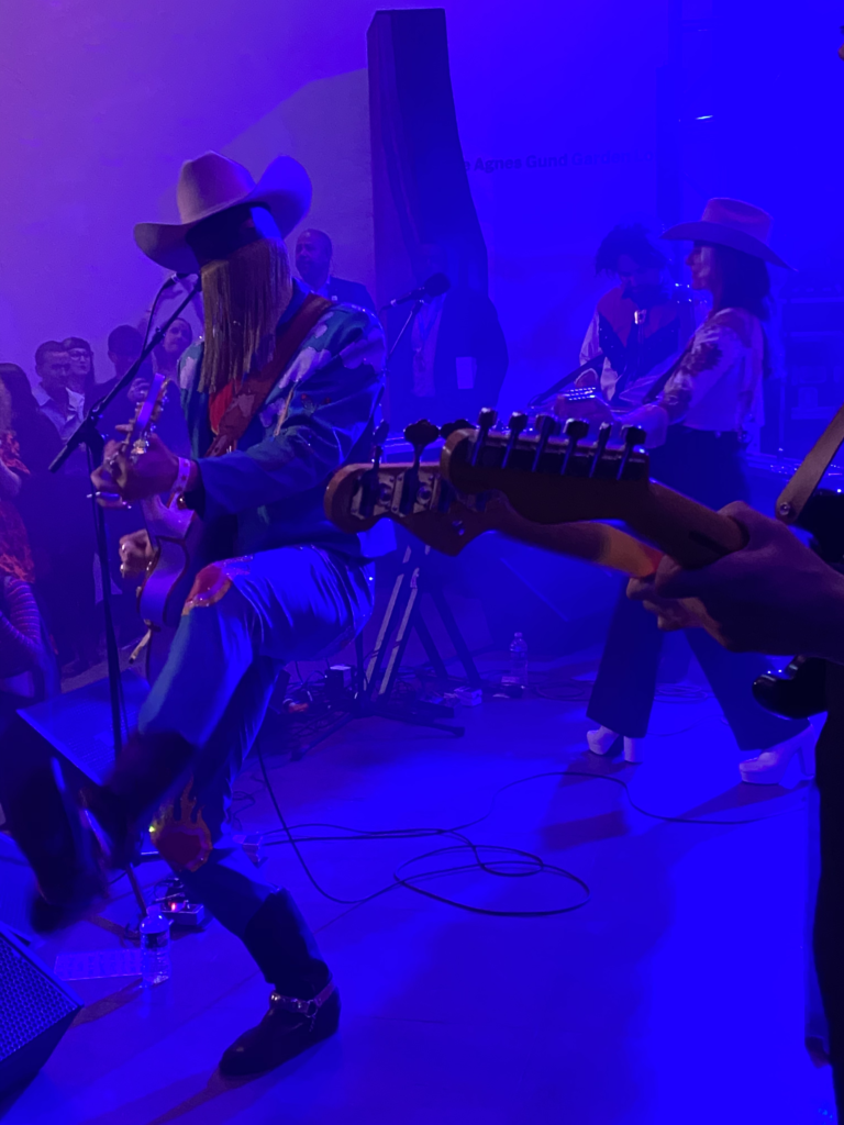 A white man in a fringed mask and a cowboy hat plays guitar surrounded by other musicians and audience members.