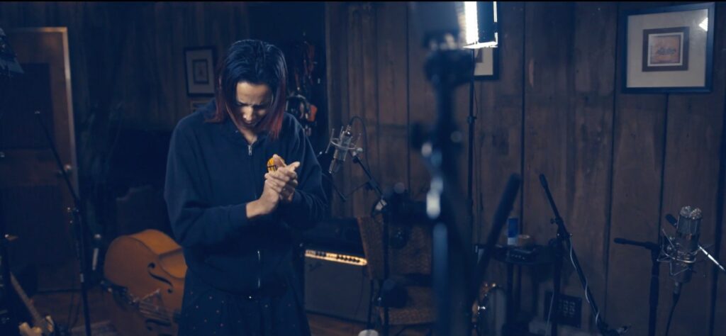 A Black woman in a sweatshirt looks down and claps among instruments and recording equipment.