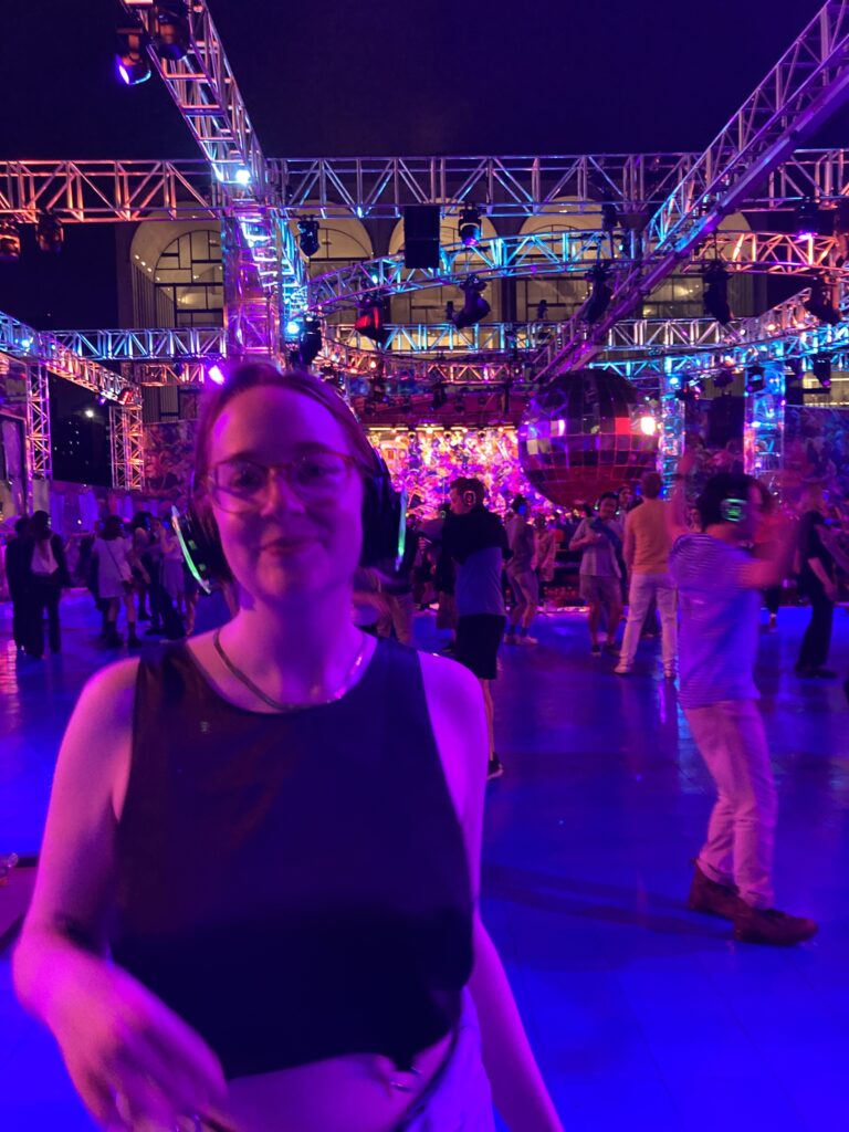 A white nonbinary person with glasses and headphones smiles in front of a large disco ball and steel structure lit with multicolored lights.