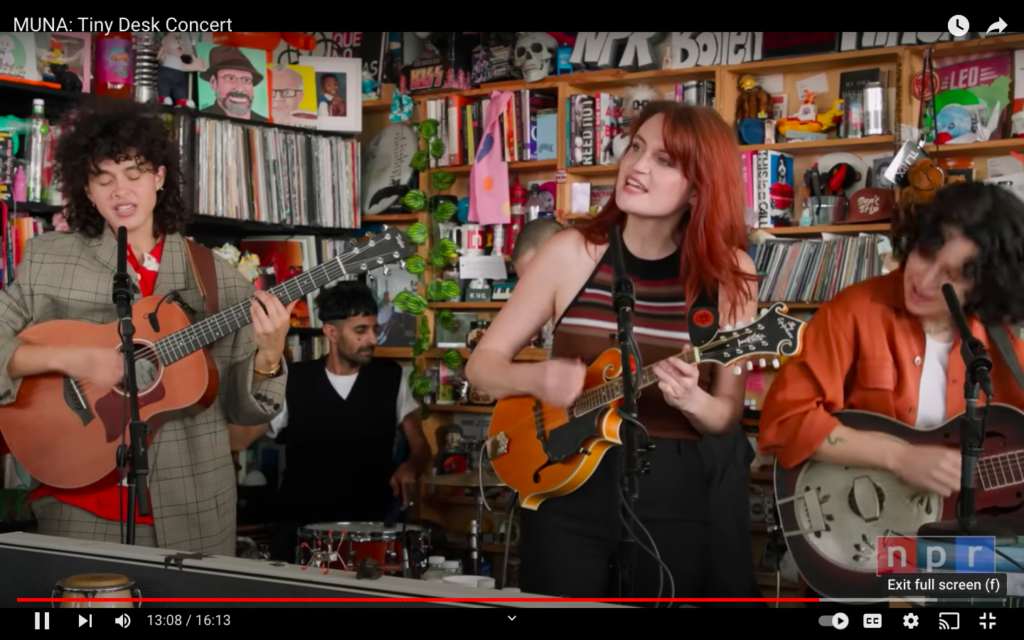 A light skinned biracial nonbinary person plays acoustic guitar next to a white redheaded woman with a mandolin and a white brunette with an electric guitar. In the background, a South Asian man plays drums.