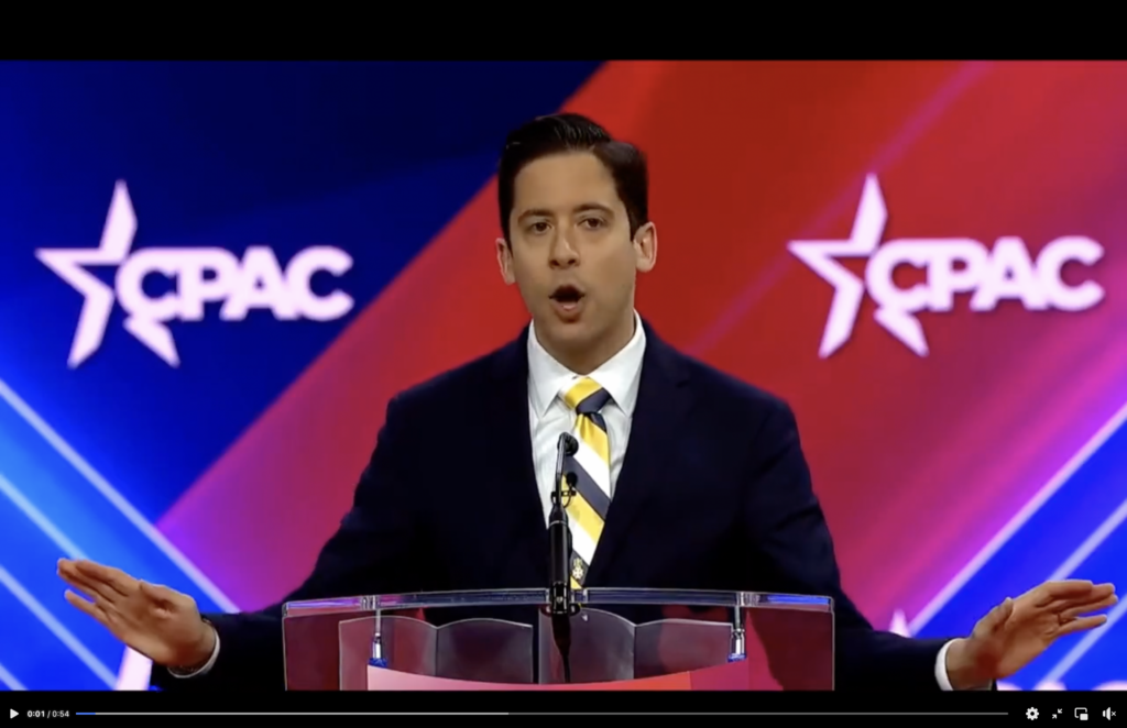 Michael Knowles stands at a lectern in front of a red, white, and blue background displaying the acronym "CPAC". 